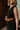 Side view of model wearing the Nova Black Sleeveless Belted Jumpsuit which features black knit fabric, upper pleated details, wide pant legs, v-neckline, sleeveless, monochrome back zipper with hook closure and an adjustable camel brown belt with a gold b