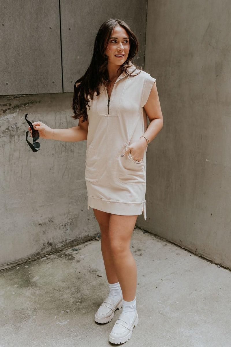 Full body front view of model wearing the Lottie Tan Quarter-Zip Sleeveless Mini Dress that has light tan knit fabric, ribbed details, two front pockets,small side slits, and a quarter zip up with a high neck.