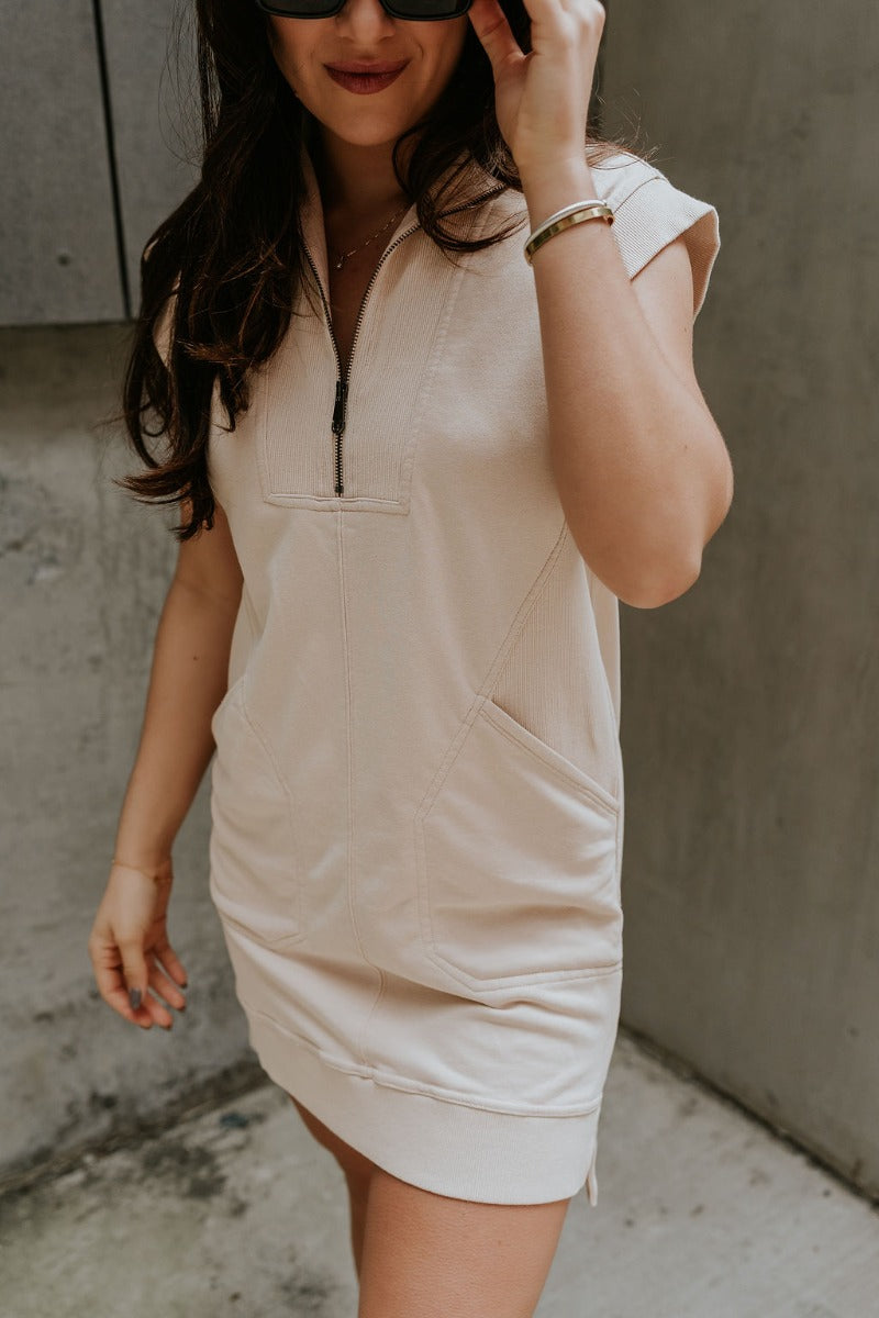 front view of model wearing the Lottie Tan Quarter-Zip Sleeveless Mini Dress that has light tan knit fabric, ribbed details, two front pockets,small side slits, and a quarter zip up with a high neck.