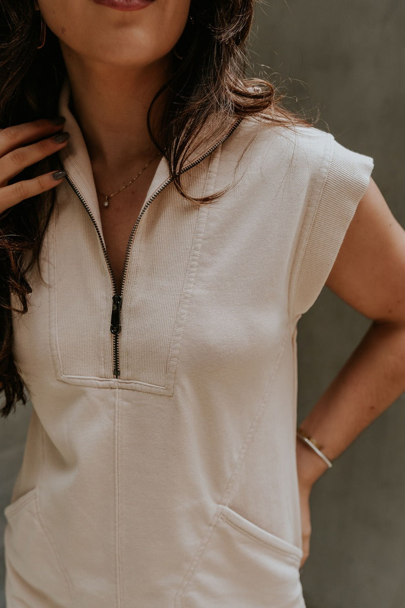 Upper front view of model wearing the Lottie Tan Quarter-Zip Sleeveless Mini Dress that has light tan knit fabric, ribbed details, two front pockets,small side slits, and a quarter zip up with a high neck.