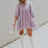 Full body front view of model wearing the Hadleigh Lavender Puff Sleeve Babydoll Dress that has lavender fabric with a plaid pattern, mini length, side pockets, a v neck with a tie closure, and half sleeves with elastic trim.