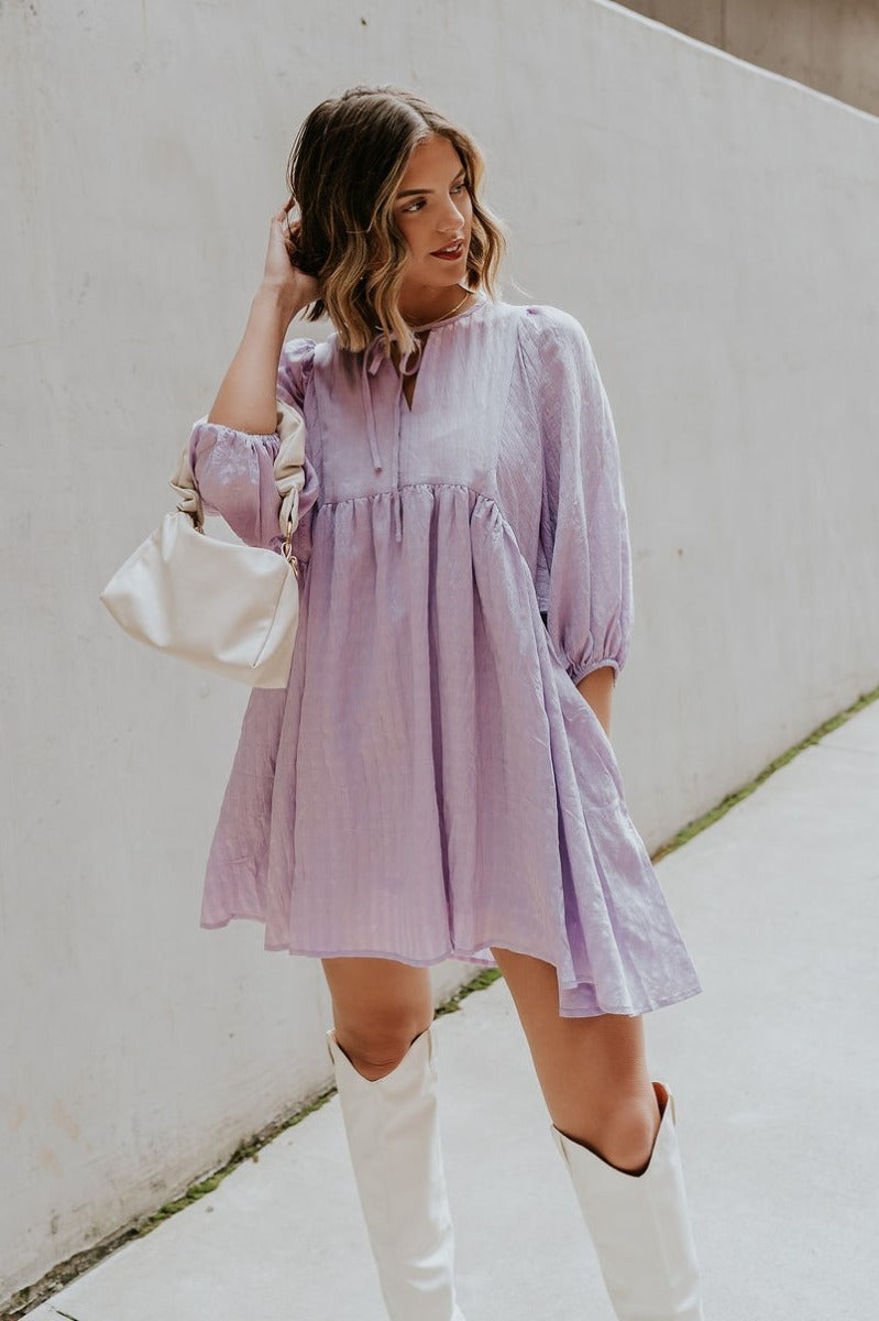 front view of model wearing the Hadleigh Lavender Puff Sleeve Babydoll Dress that has lavender fabric with a plaid pattern, mini length, side pockets, a v neck with a tie closure, and half sleeves with elastic trim.