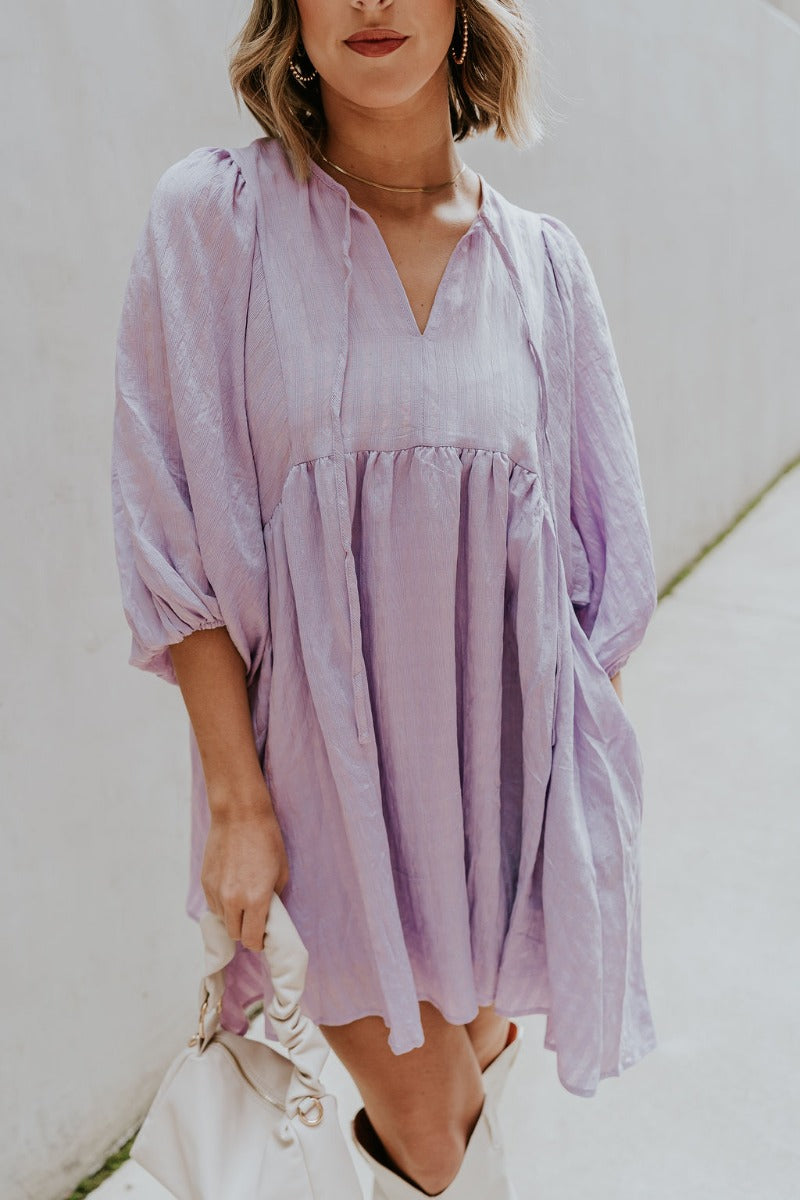 Close front view of model wearing the Hadleigh Lavender Puff Sleeve Babydoll Dress that has lavender fabric with a plaid pattern, mini length, side pockets, a v neck with a tie closure, and half sleeves with elastic trim.