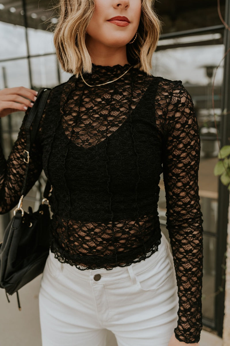 Close front view of model wearing the Abby Black Lace Long Sleeve Top that has black sheer fabric, monochrome lace floral pattern, lettuce hem details, a high neck and long sleeves.