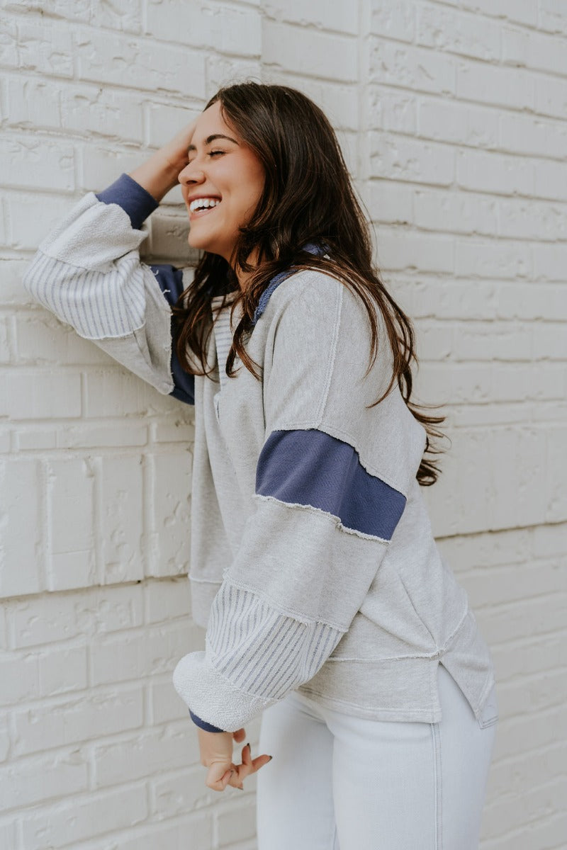 Side view of model wearing the Rylan Grey & Navy Color Block Sweatshirt that has navy blue and heather grey knit fabric, color block striped pattern, raw hems, v-neck with collar, and long sleeves with cuffs.
