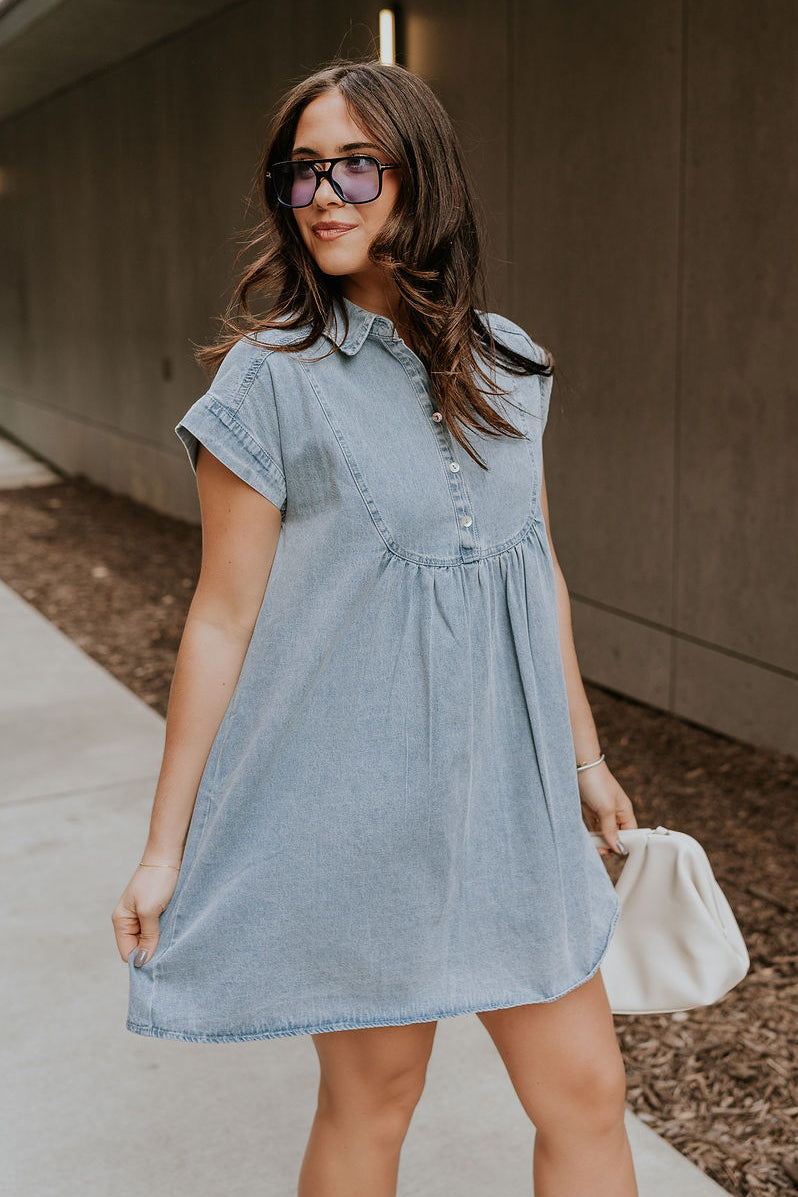 Side view of model wearing the Everly Denim Short Sleeve Buttoned Dress which features light denim wash fabric, mini length, two front slit pockets, quarter pearlescent button up, collared neckline and short sleeves.