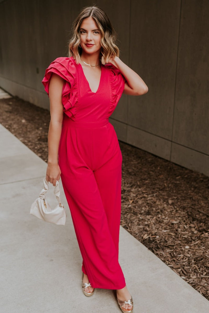 Full body front view of model wearing the Giana Hot Pink Wide Leg Ruffle Jumpsuit that has hot pink knit fabric, pockets, a ruffle details, a v-neck, a back zipper, and flare pant legs.