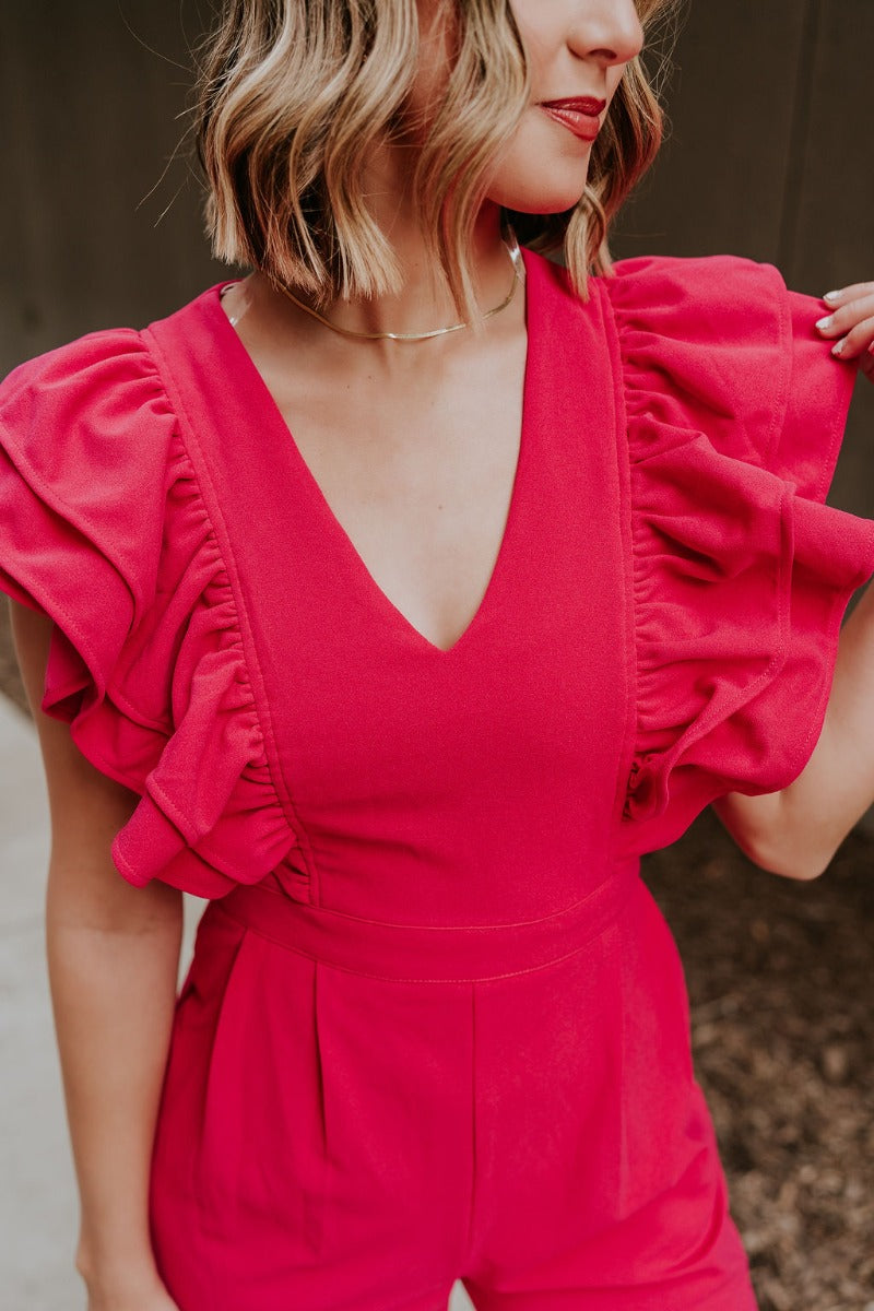 Upper front view of model wearing the Giana Hot Pink Wide Leg Ruffle Jumpsuit that has hot pink knit fabric, pockets, a ruffle details, a v-neck, a back zipper, and flare pant legs.