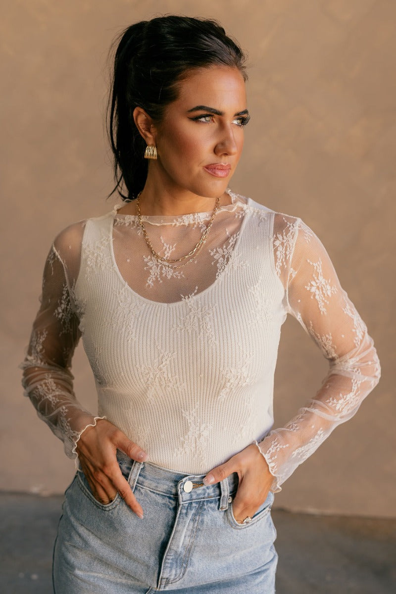 Front view of model wearing the Nadia Cream Lace Long Sleeve Top which features cream sheer fabric, lettuce hem details, lace floral design, high neckline and long sleeves.