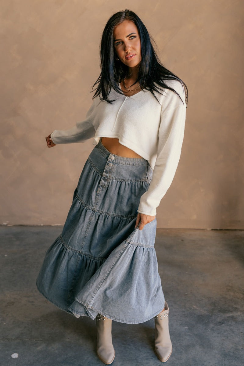 Full body view of model wearing the Alice Light Denim Tiered Midi Skirt which features light denim fabric, three tiered design, midi length, two slit pockets, beige stitching and front button up closure.