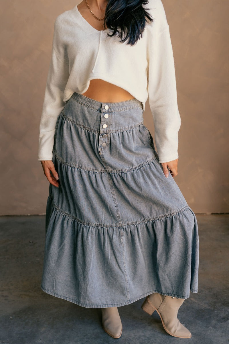 Front view of model wearing the Alice Light Denim Tiered Midi Skirt which features light denim fabric, three tiered design, midi length, two slit pockets, beige stitching and front button up closure.