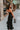 Full body back view of model wearing the Colette Black Floral Satin Midi Dress that has black satin fabric, monochrome florals, a assymetrical ruffle tier design, pleated fabric, a cowl neck, and thin straps.