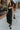 Full body front view of model wearing the Colette Black Floral Satin Midi Dress that has black satin fabric, monochrome florals, a assymetrical ruffle tier design, pleated fabric, a cowl neck, and thin straps.