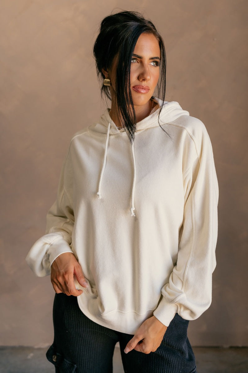 Front view of model wearing the Charlie Cream Drawstring Hoodie Sweatshirt which features cream knit fabric, ribbed thick hem, exposed seam details, a high neckline, a hood with drawstrings, and wide long sleeves with ribbed cuffs.