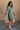 Full body side view of model wearing the Sasha Grey Green Mock Neck Sweater Dress which features grey green textured knit fabric, a thick ribbed hem, mini length, a high ribbed neckline, and long sleeves with ribbed cuffs.