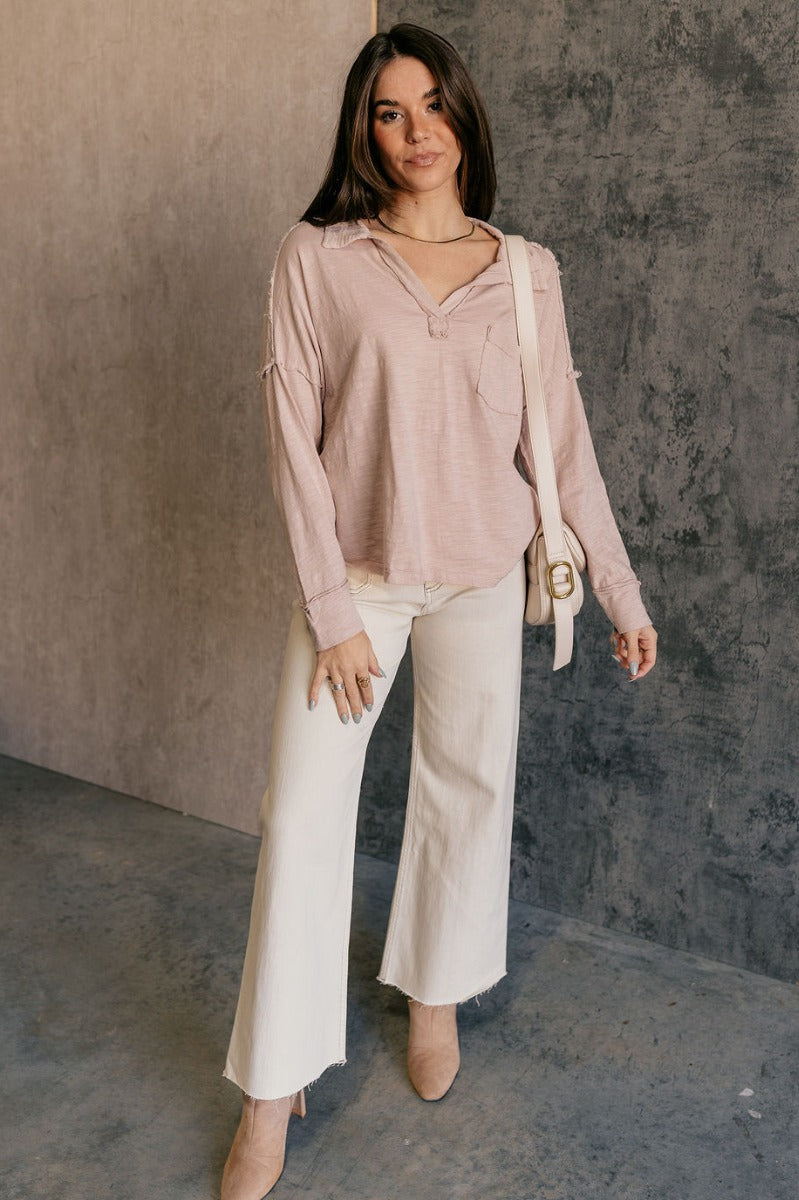 Full body view of model wearing the Ada Dusty Rose Long Sleeve Top which features dusty mauve pink knit fabric, slight slits on each side, distressed details, a front left chest pocket, a v-neckline with collar, dropped shoulders, and long sleeves with cu