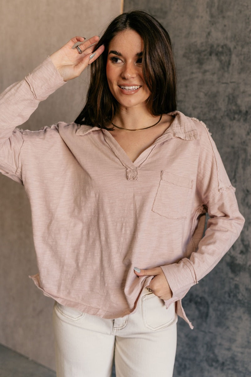 Front view of model wearing the Ada Dusty Rose Long Sleeve Top which features dusty mauve pink knit fabric, slight slits on each side, distressed details, a front left chest pocket, a v-neckline with collar, dropped shoulders, and long sleeves with cuffs.