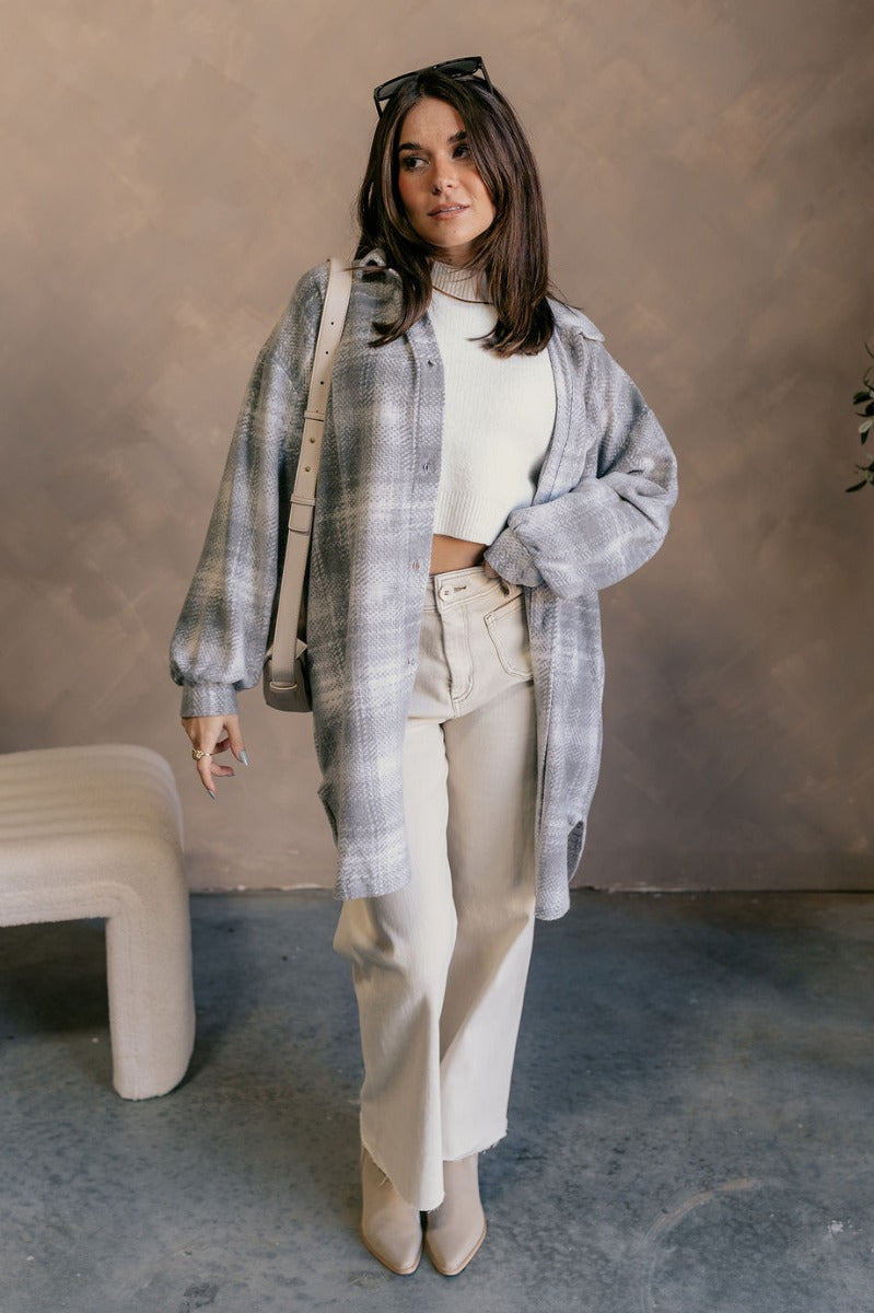 Full body view of model wearing the Clara Grey Plaid Knit Long Sleeve Shacket which features grey and white knit fabric, a geometric plaid pattern, mid thigh length hem, two pockets on each side, tortoise buttons, a collared neckline, a left front chest p