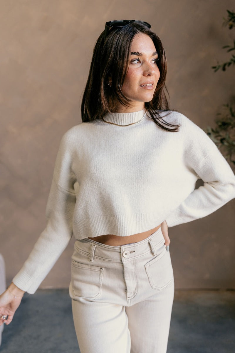 Front view of model wearing the Emily Ivory Cropped Long Sleeve Sweater which features ivory knit fabric, a ribbed hem, a cropped waist, a slight high neckline, dropped shoulders, and long sleeves.