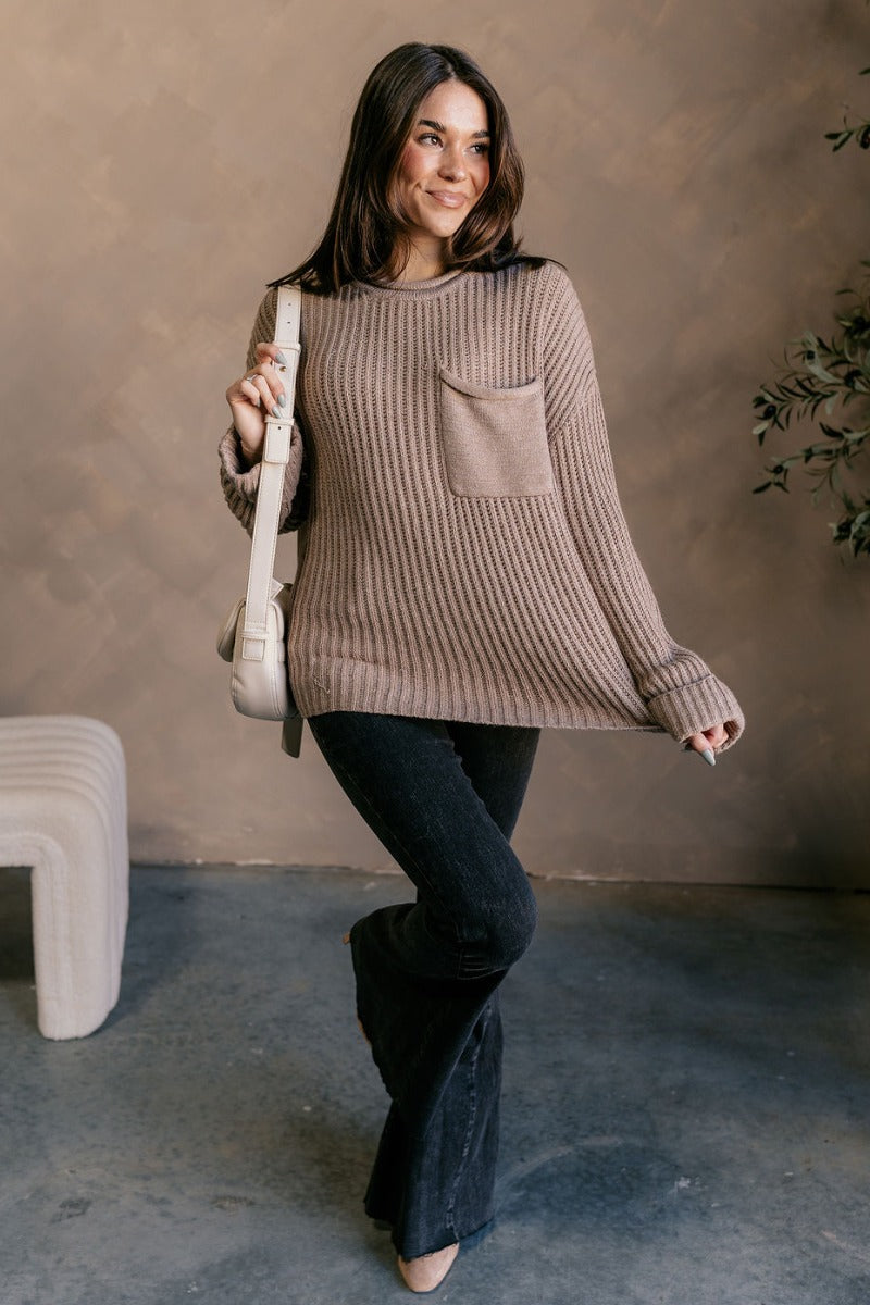 Full body view of model wearing the Mila Mocha Cable Knit Long Sleeve Sweater which features mocha cable knit fabric, a front left chest pocket, a round neckline, dropped shoulders, and long sleeves with folded cuffs.