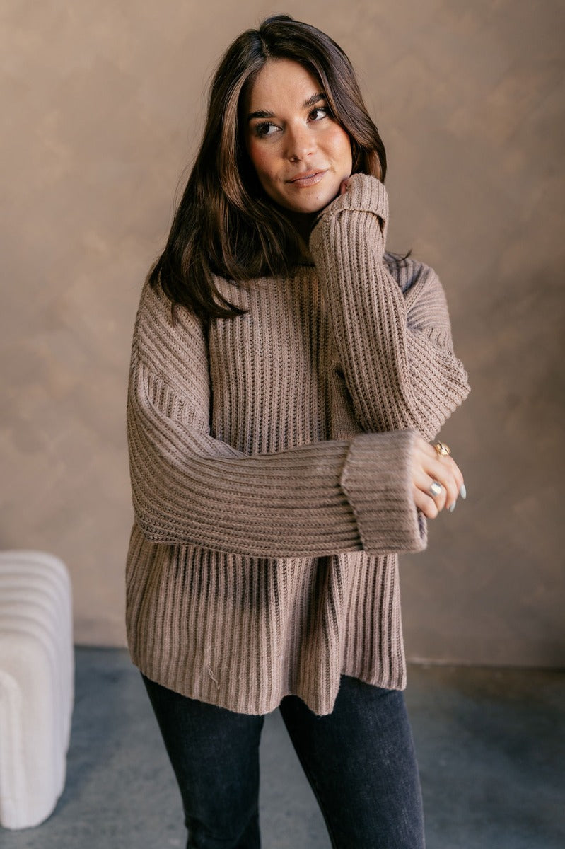 Front view of model wearing the Mila Mocha Cable Knit Long Sleeve Sweater which features mocha cable knit fabric, a front left chest pocket, a round neckline, dropped shoulders, and long sleeves with folded cuffs.