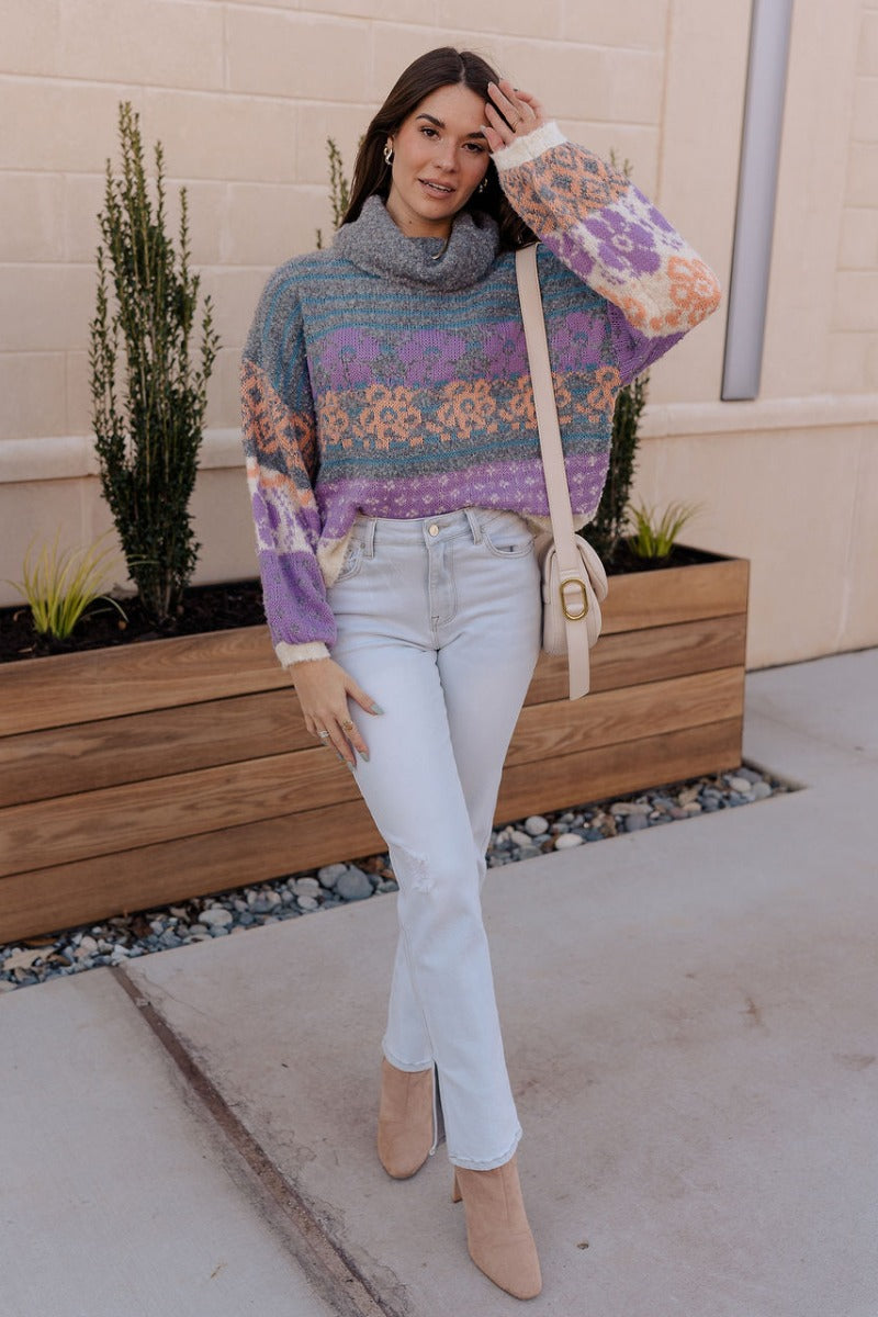 Full body view of model wearing the Eliza Purple Multi Long Sleeve Sweater which features purple, peach, cream blue and teal grey knit fabric, a floral and striped pattern, a loose turtle neckline, dropped shoulders, and long balloon sleeves with ribbed c