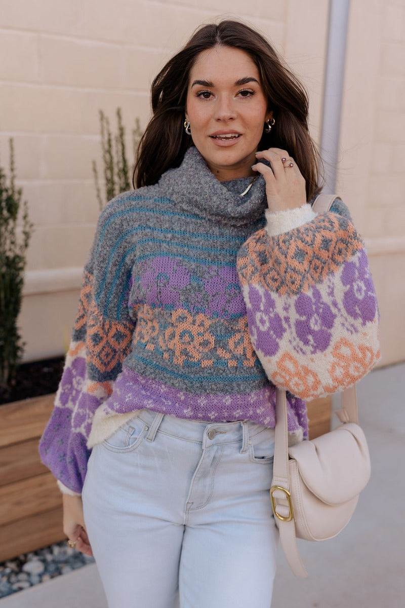 Front view of model wearing the Eliza Purple Multi Long Sleeve Sweater which features purple, peach, cream blue and teal grey knit fabric, a floral and striped pattern, a loose turtle neckline, dropped shoulders, and long balloon sleeves with ribbed cuffs