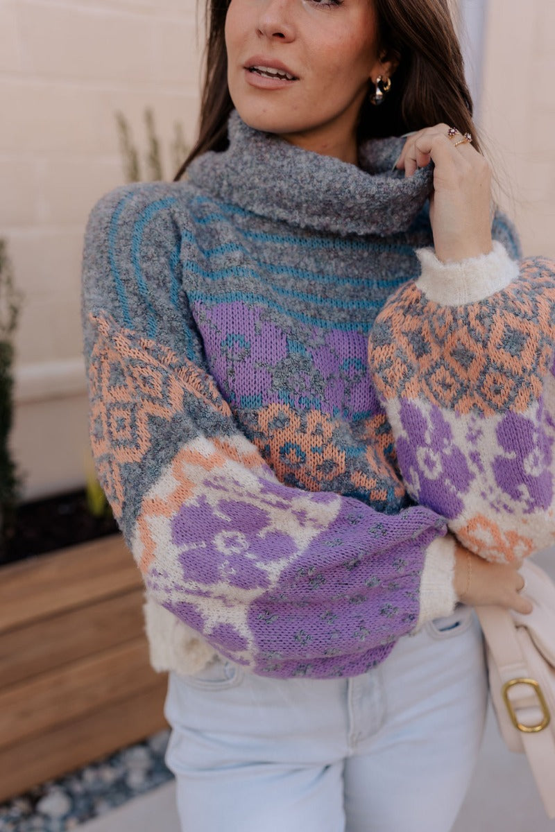 Close up view of model wearing the Eliza Purple Multi Long Sleeve Sweater which features purple, peach, cream blue and teal grey knit fabric, a floral and striped pattern, a loose turtle neckline, dropped shoulders, and long balloon sleeves with ribbed cu