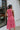 Back view of model wearing the Lost Without You Midi Dress that has a red and pink geometric pattern, a tiered skirt with a midi-length hem, and a draped neck that ties in the back.
