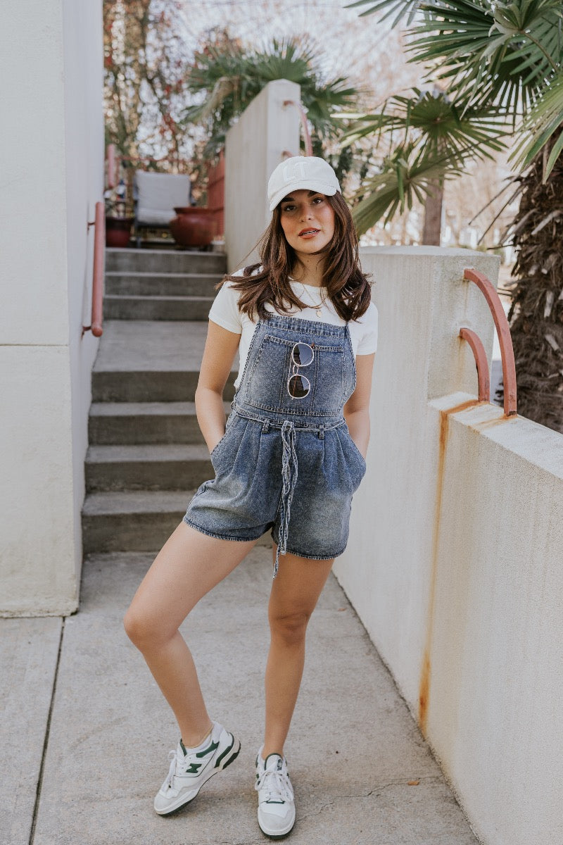 Front image of model wearing The Marina Washed Denim Romper features medium wash denim fabric, two front pockets, two back pockets, belt loops with a drawstring tie, one front chest pocket, a halter neckline, adjustable tie straps, a side zipper closure a