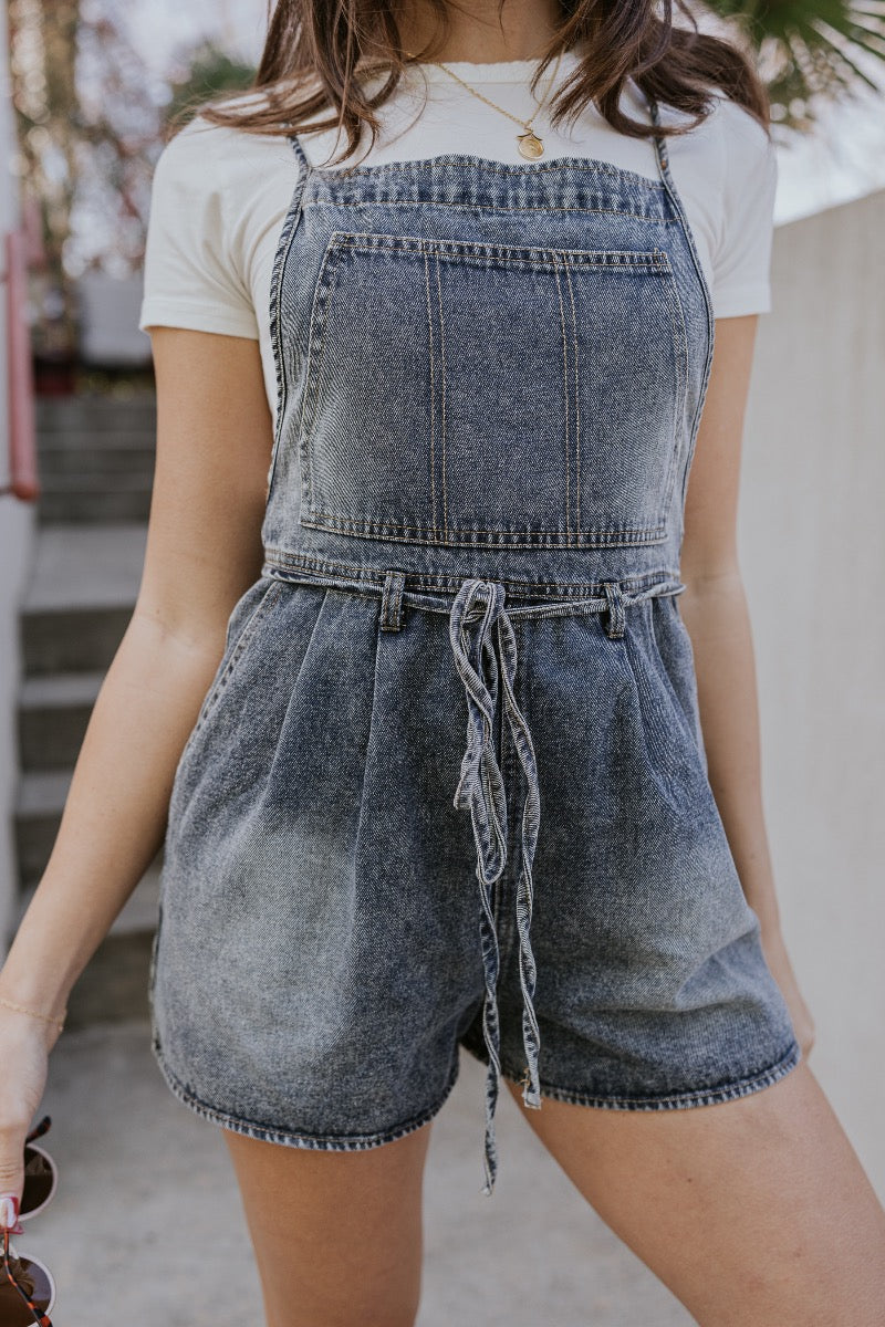 Front detail image of model wearing The Marina Washed Denim Romper features medium wash denim fabric, two front pockets, two back pockets, belt loops with a drawstring tie, one front chest pocket, a halter neckline, adjustable tie straps, a side zipper cl