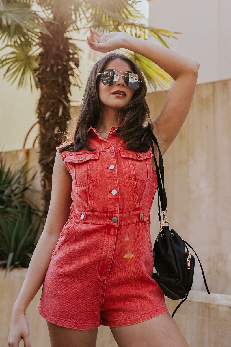 front image of modeal wearing The Emily Washed Red Denim Sleeveless Romper features washed red denim fabric, two front pockets, silver button details, button-up front, two front chest buttoned pockets, two back pockets, a collared neckline and a sleeveles