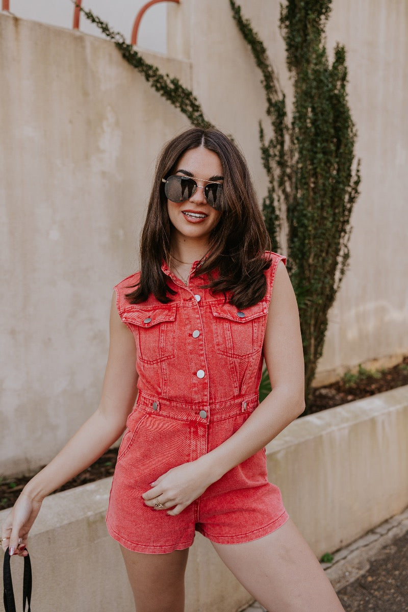 front detail image of modeal wearing The Emily Washed Red Denim Sleeveless Romper features washed red denim fabric, two front pockets, silver button details, button-up front, two front chest buttoned pockets, two back pockets, a collared neckline and a sl