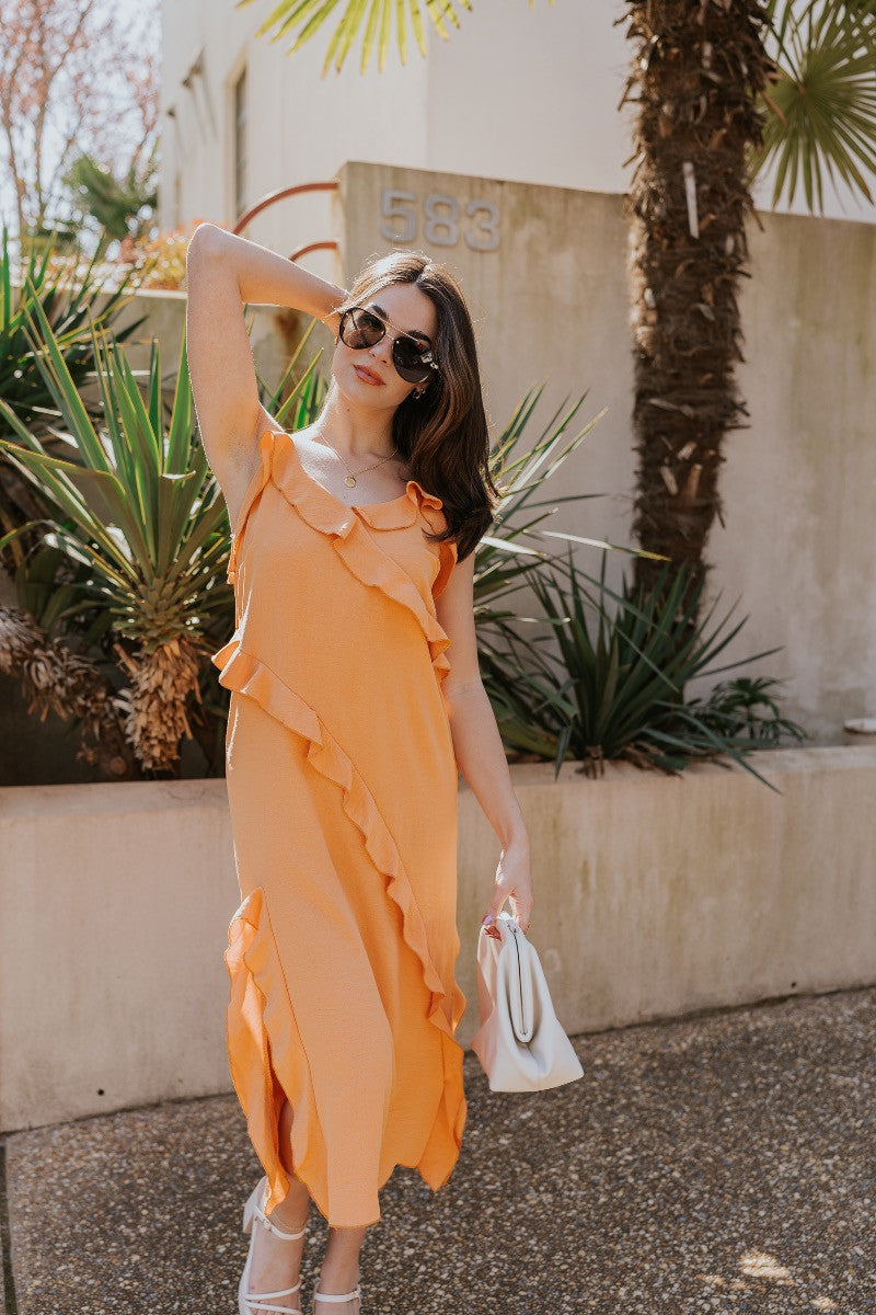 Full body view of model wearing the Serena Orange Ruffle Midi Dress which features orange light weight fabric, midi length, ruffle hem with slight slit detail, ruffle tiered design, sweetheart neckline, adjustable straps and sleeveless.