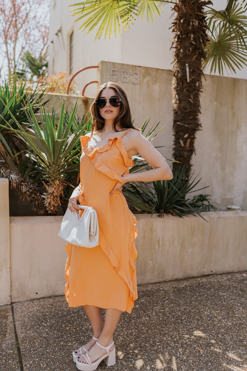 Full body side view of model wearing the Serena Orange Ruffle Midi Dress which features orange light weight fabric, midi length, ruffle hem with slight slit detail, ruffle tiered design, sweetheart neckline, adjustable straps and sleeveless.