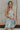 Front view of model wearing the Head Over Heels Floral Dress that has cream, blue, purple, green and light pink sheer floral fabric, a babydoll style, a square neck, spaghetti straps, an open back with a tied bow, and a back zipper closure