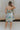 Full body back view of model wearing the Head Over Heels Floral Dress that has cream, blue, purple, green and light pink sheer floral fabric, a babydoll style, a square neck, spaghetti straps, an open back with a tied bow, and a back zipper closure