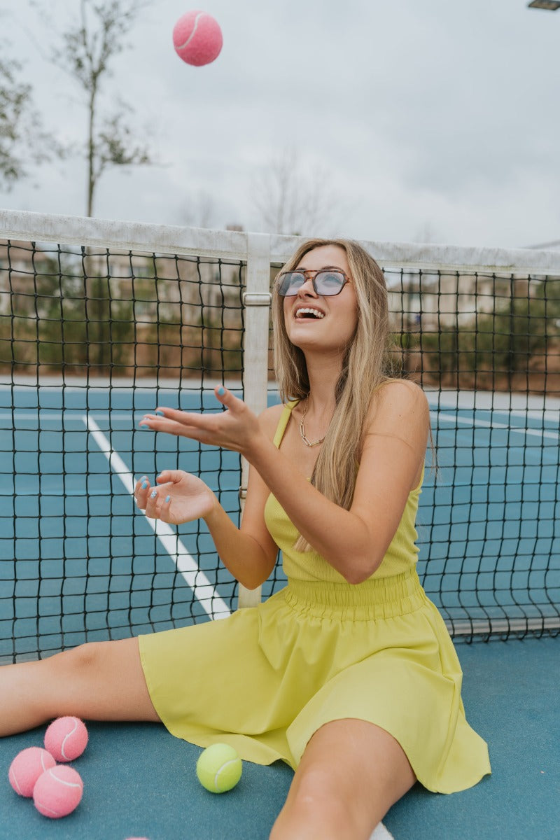 Image of model wearing sitting on a tennis court while wearing the  Movement Mini Dress in Lime.