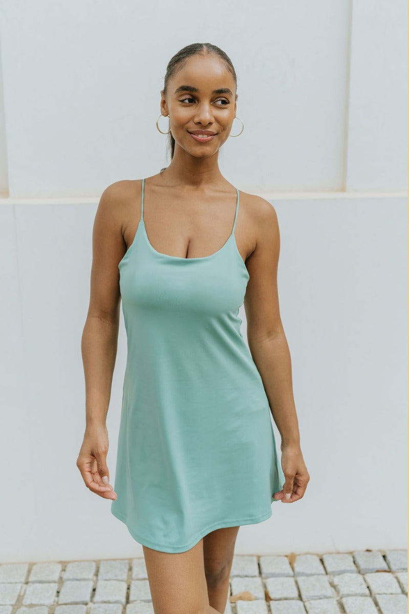 Front view of model wearing the Traveler Mini Dress in Sage that has sage green fabric, shorts lining with side pockets, bra padding, a scoop  neck, and straps that cross in the back