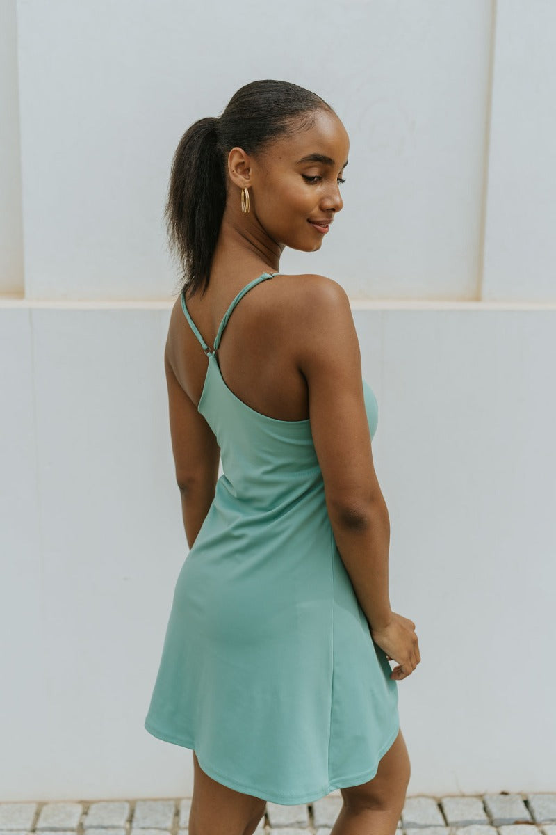 Side view of model wearing the Traveler Mini Dress in Sage that has sage green fabric, shorts lining with side pockets, bra padding, a scoop  neck, and straps that cross in the back