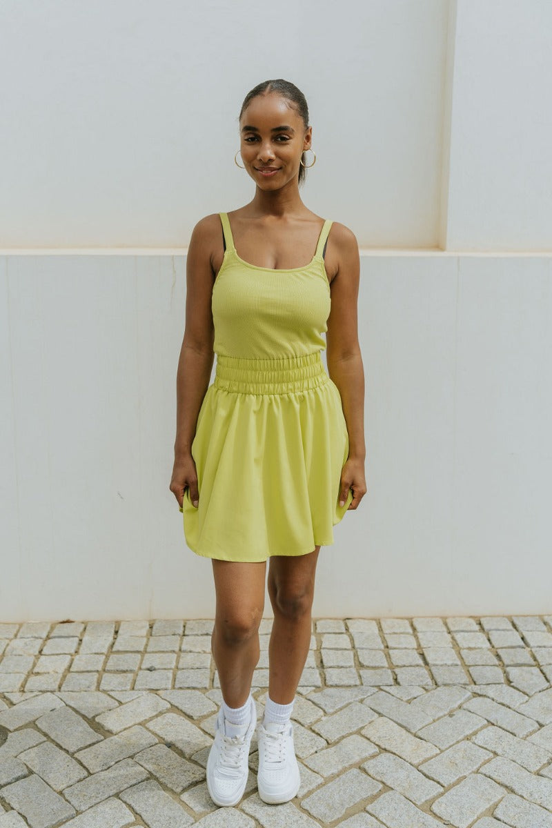 Full body front view of model wearing the Movement Mini Dress in Lime that has a yellow-green ribbed upper body, ribbed shorts lining, a lightweight skirt, a smocked waist, a scoop neck, and thick straps