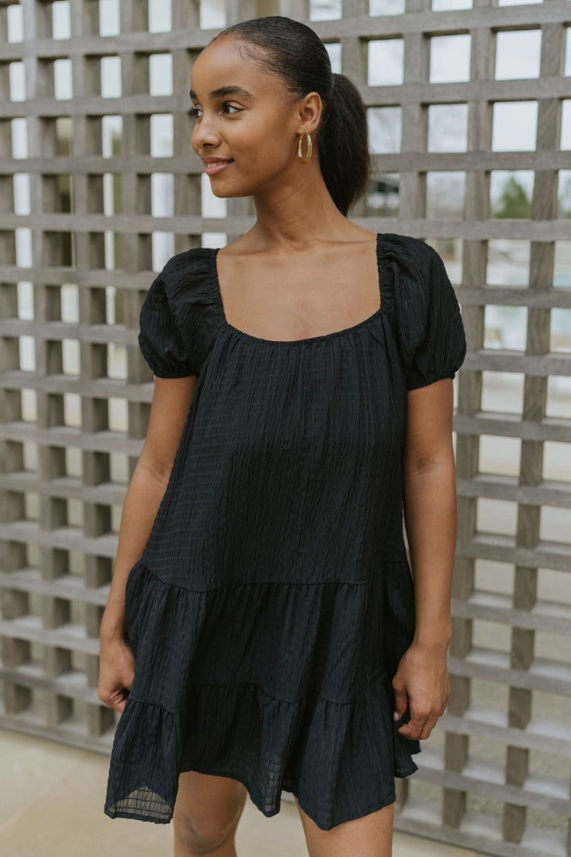 Front view of model wearing the Change Your Mind Dress that has black textured fabric, mini length, a ruffle hem, a square neckline, short puff sleeves with elastic bands, a back tie strap, and black lining.