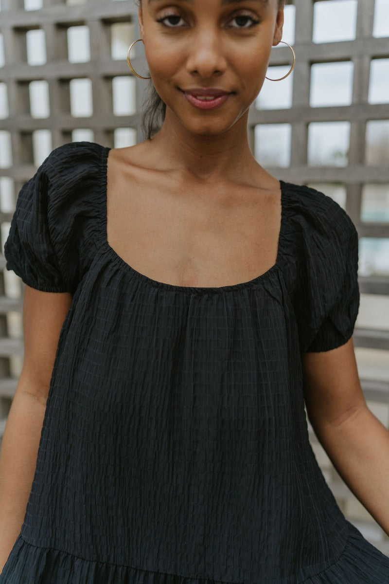Close upper front view of model wearing the Change Your Mind Dress that has black textured fabric, mini length, a ruffle hem, a square neckline, short puff sleeves with elastic bands, a back tie strap, and black lining.