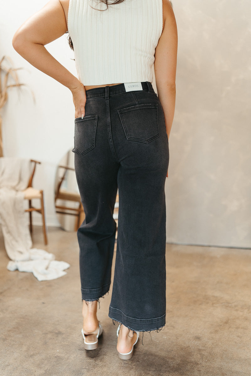 Back view of model wearing the Ceros: Florence Wide Leg Jeans that have black denim, a zipper and button closure, belt loops, high-rise waist, pockets, and wide cropped legs with distressed hems