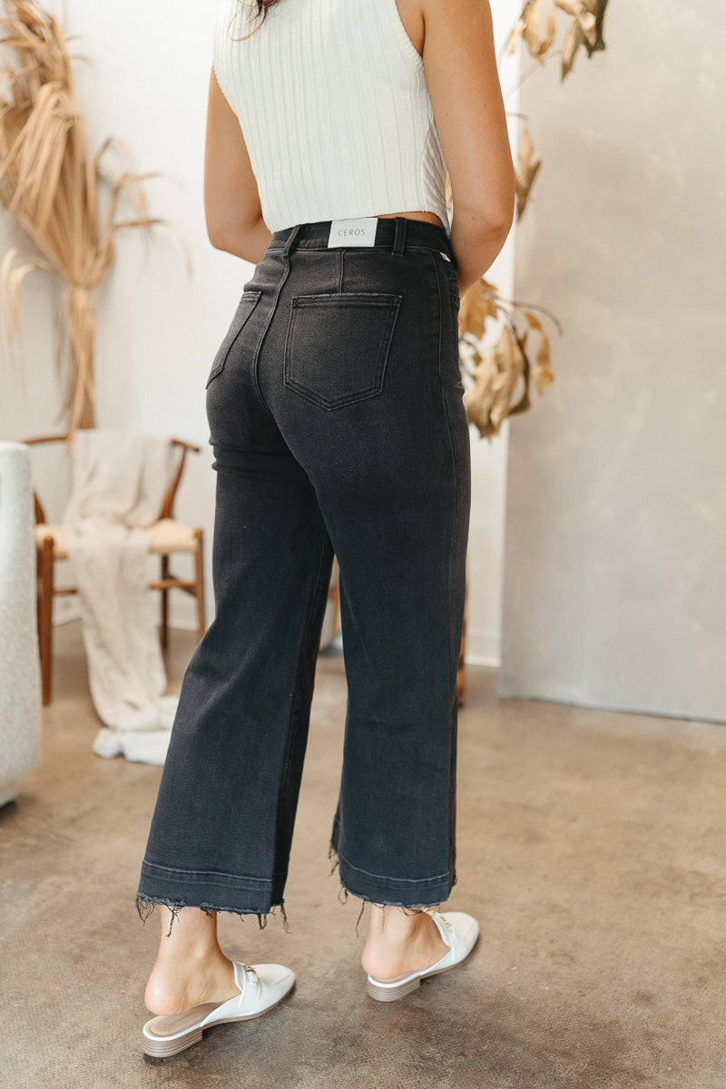 Back view of model wearing the Ceros: Florence Wide Leg Jeans that have black denim, a zipper and button closure, belt loops, high-rise waist, pockets, and wide cropped legs with distressed hems