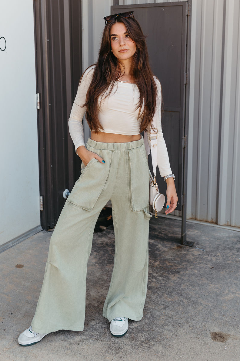 Full body view of model wearing the Ellison Denim Cargo Pants in Light Sage which features light olive cotton fabric, two front pockets, a side cargo loop detail, an elastic waistband and wide legs.