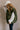 Side view of model wearing the Big Sky Olive Green Oversized Puffer Vest which features olive green lightweight puffer fabric, an elastic hem, two front pockets, a monochromatic front zipper, a high neckline, and a sleeveless design.