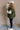 Full body side view of model wearing the Big Sky Olive Green Oversized Puffer Vest which features olive green lightweight puffer fabric, an elastic hem, two front pockets, a monochromatic front zipper, a high neckline, and a sleeveless design.
