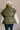 Back view of model wearing the Big Sky Olive Green Oversized Puffer Vest which features olive green lightweight puffer fabric, an elastic hem, two front pockets, a monochromatic front zipper, a high neckline, and a sleeveless design.