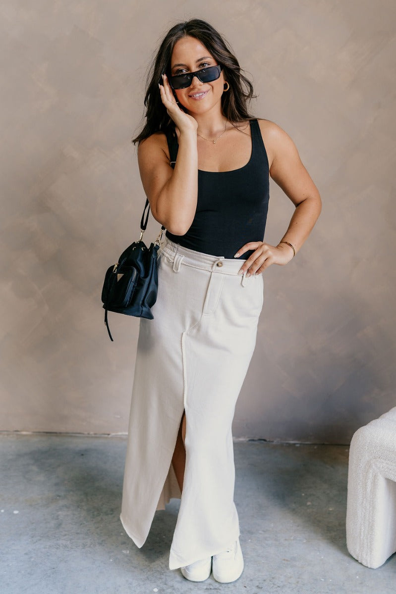 Full body view of model wearing the Tori Cream Front Slit Midi Skirt which features cream fabric, a front slit, a tortoise button closure, a back elastic waistband, belt loops, front pockets, distressed details in the back, and a raw hem.